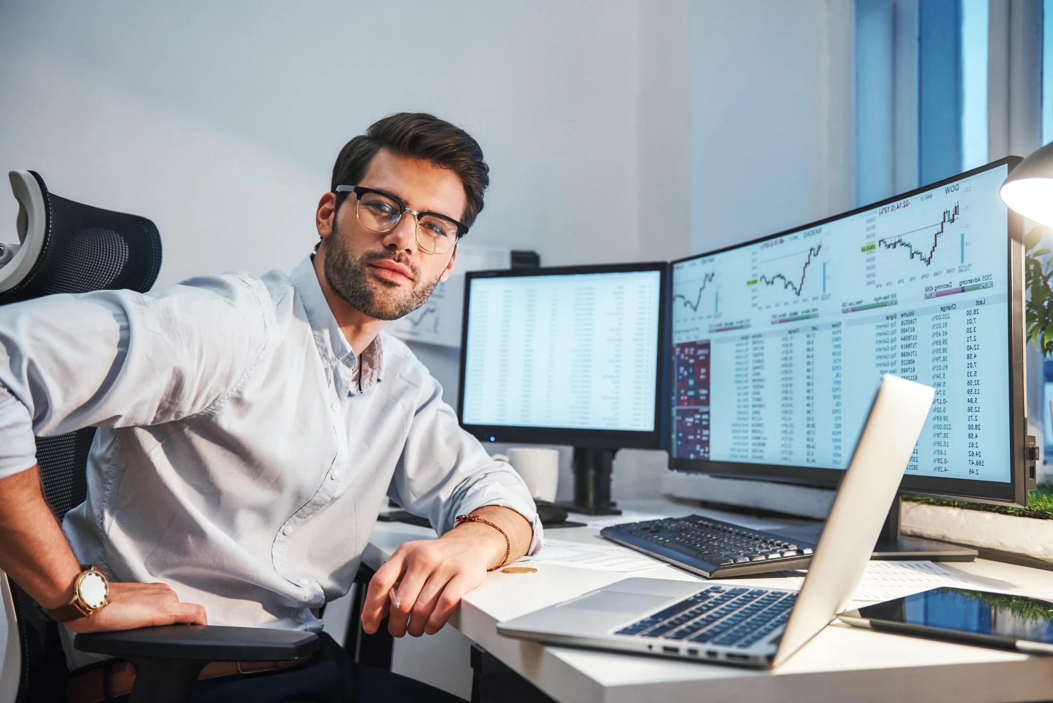 Confident and smart. Young trader or businessman in eyewear and formal clothes is looking at camera while sitting in front of computer screens with financial data in his modern office.